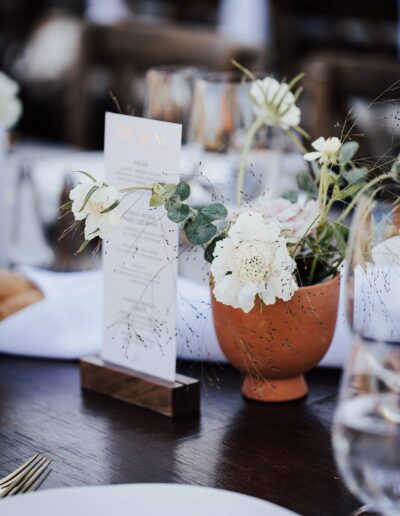 A table setting featuring a small pot of white flowers and greenery, a menu held upright in a wooden holder, and a place setting with a glass and plate.