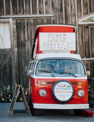 A red and white ice cream truck with a rooftop sign that says "ICE CREAM," parked in front of a wooden building. A chalkboard menu stands on the ground beside the truck.
