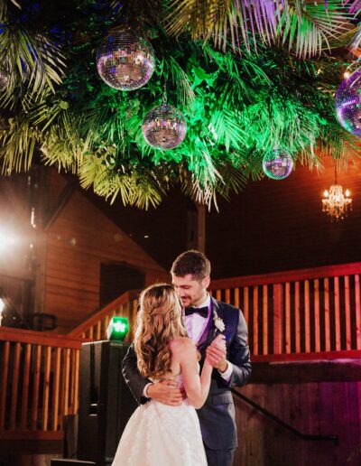 A couple in formal attire dances under hanging disco balls and greenery, with wooden railings and soft lighting in the background.
