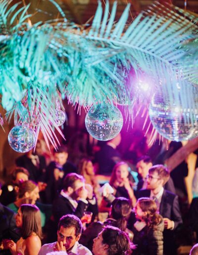 People socializing under hanging palm leaves and disco balls in a vibrant, colorful indoor setting.