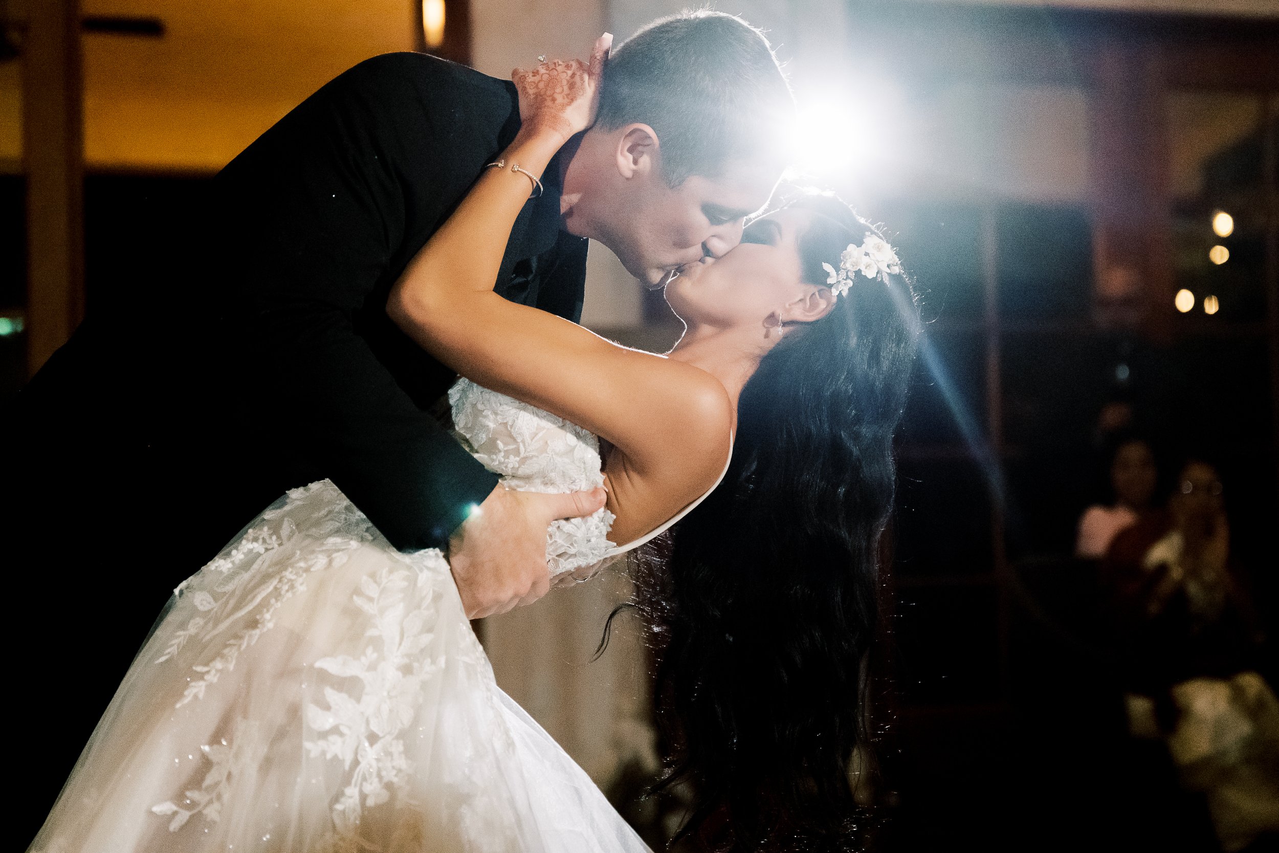 A bride and groom share a kiss while dancing, with the groom dipping the bride backward. The bride wears a lace wedding gown, and the groom wears a black suit. A bright light shines behind them.