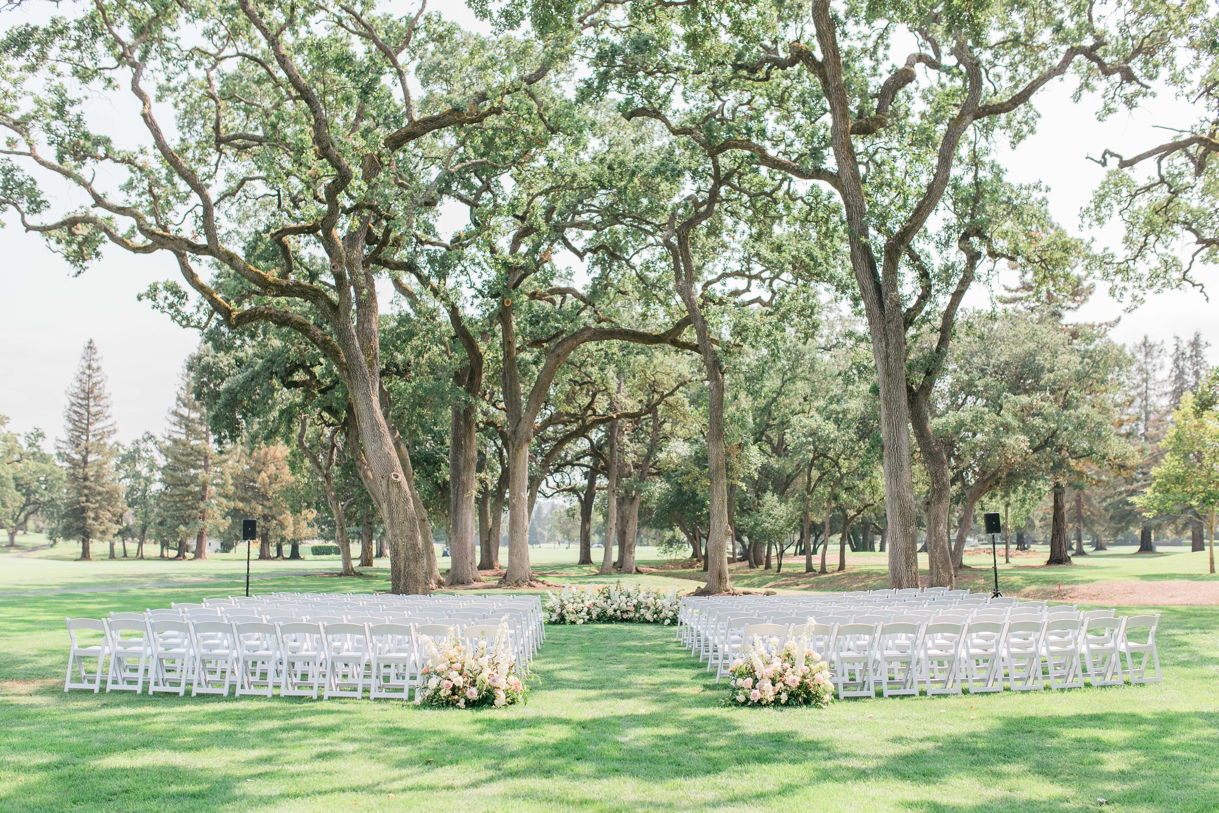 Rows of white chairs arranged outdoors on a grassy area beneath large trees, set up for a wedding ceremony with floral arrangements at the front.