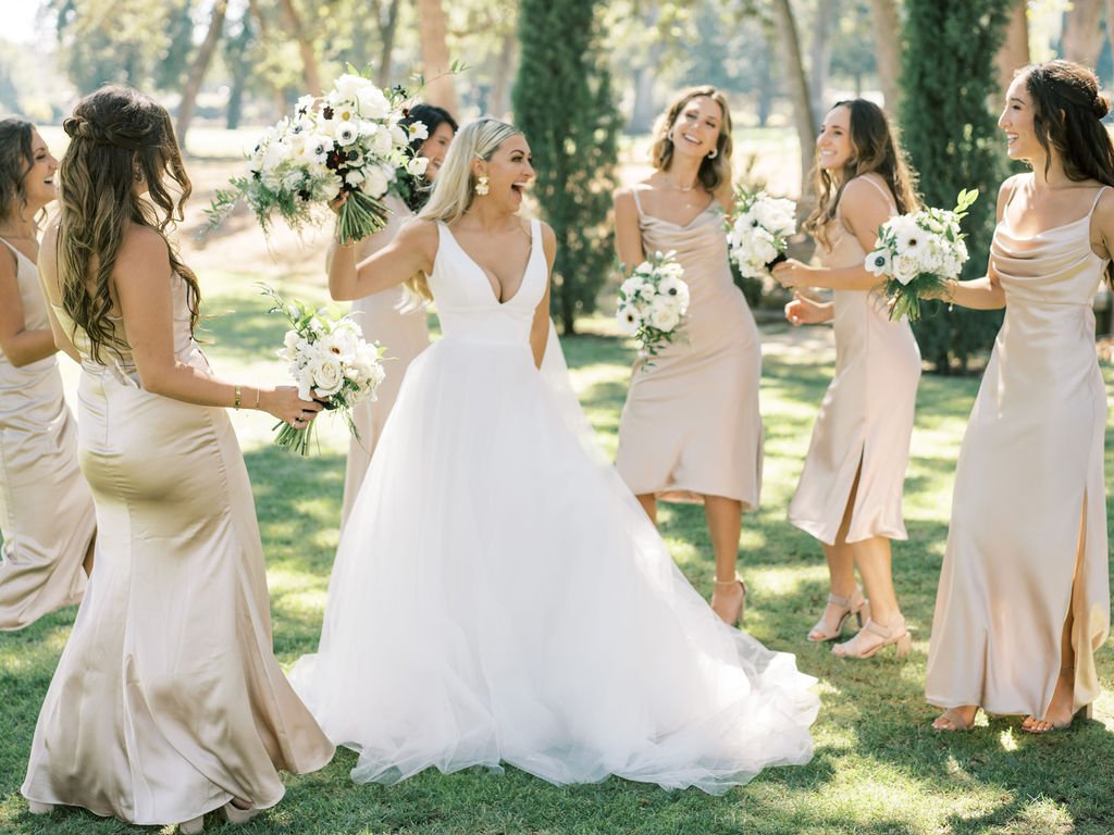 A bride in a white gown and her bridesmaids in light beige dresses holding bouquets stand together outside on a sunny day, smiling and celebrating.