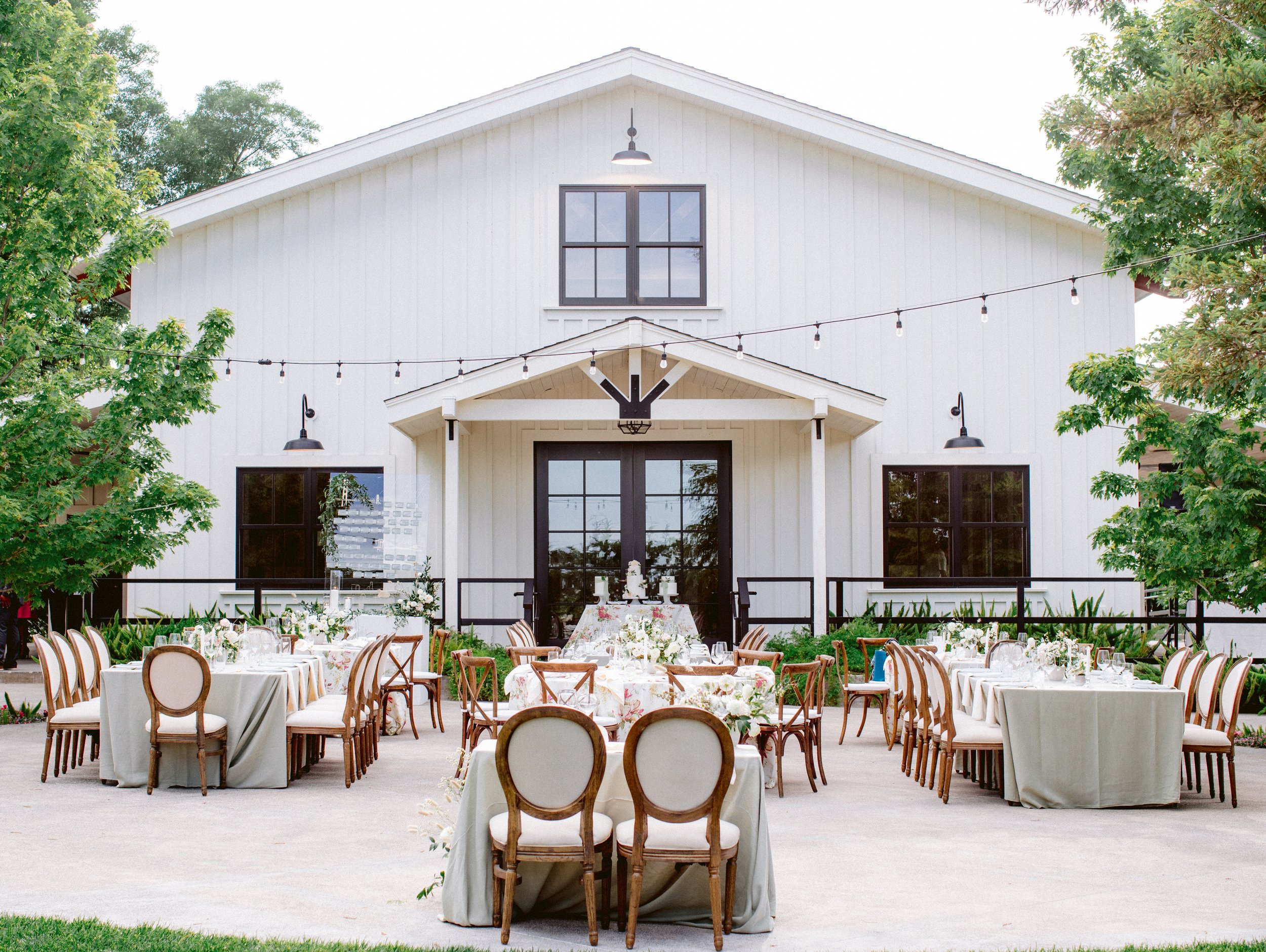 An outdoor event setup with tables and chairs is arranged in front of a large white barn-style building. String lights are hung overhead, and the area is surrounded by greenery.