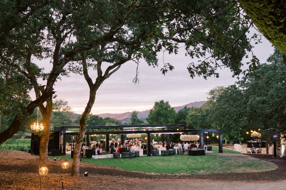 An outdoor event with people gathered under a canopy structure with chandeliers, set amidst trees and hills in the background at dusk.