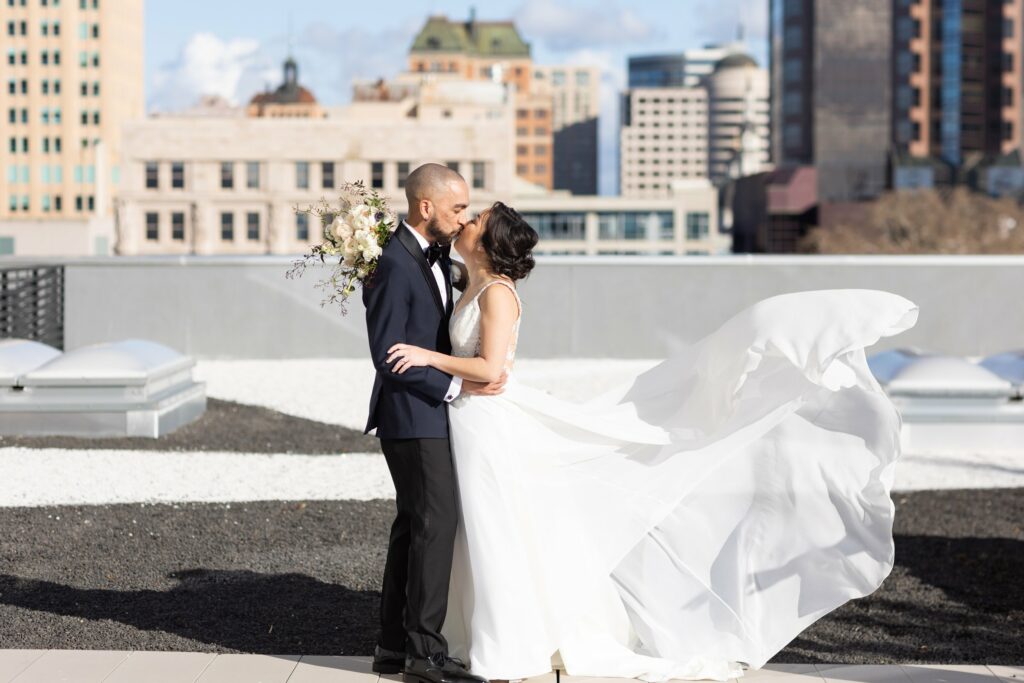 A couple in wedding attire shares a kiss on a rooftop with a cityscape in the background. The bride's dress flows in the wind.