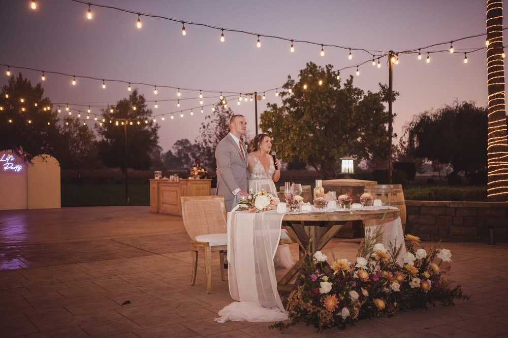 A couple stands behind a wooden table adorned with floral arrangements at an outdoor wedding reception decorated with string lights and a neon sign that reads "Let's Party".