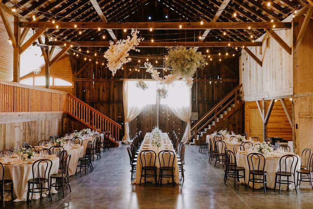 A rustic barn decorated for an event with round and rectangular tables set with white tablecloths and chairs, overhead string lights, and floral arrangements suspended from the ceiling.