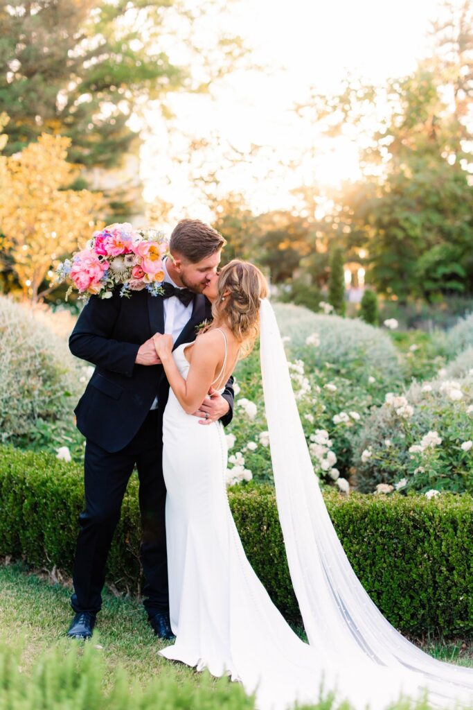 A bride and groom share a kiss in a garden setting; the groom in a black suit holds a bouquet, and the bride in a white dress has a long veil trailing behind her.