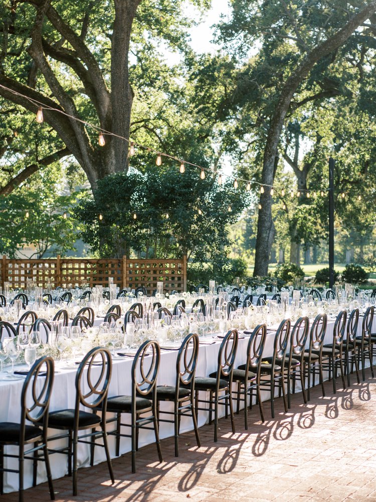 Long tables with white tablecloths and black chairs set for an outdoor event beneath trees with string lights overhead.