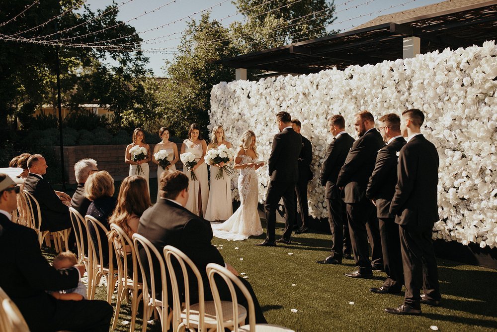 Outdoor wedding ceremony with a bride and groom standing before a white floral backdrop. Bridesmaids in white dresses and groomsmen in black suits stand on either side. Guests are seated.