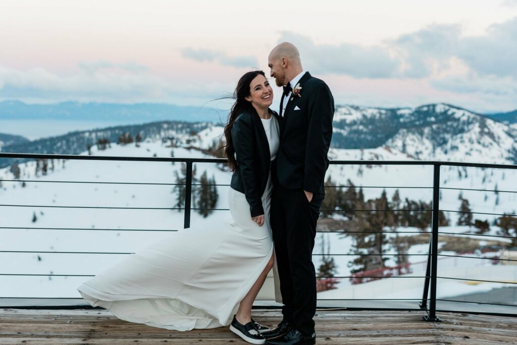 A couple, dressed in wedding attire, stand on a snowy mountain overlook. The woman in a white dress and jacket smiles, while the man in a suit kisses her head. Snow-covered mountains are in the background.