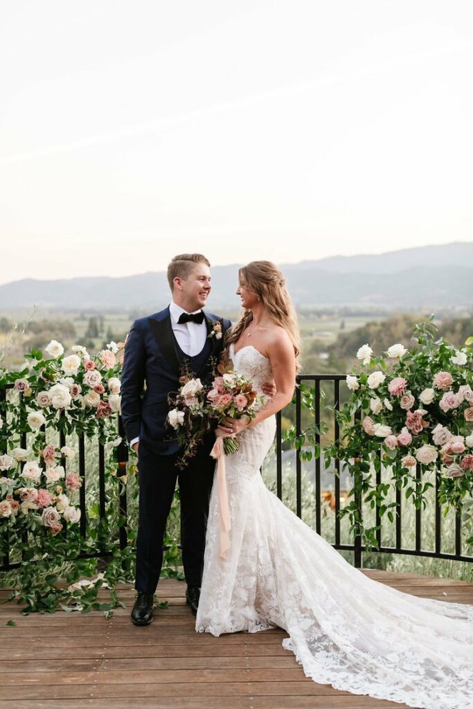 A couple in wedding attire stands on a wooden deck adorned with floral arrangements. They look at each other lovingly with a scenic landscape visible in the background.