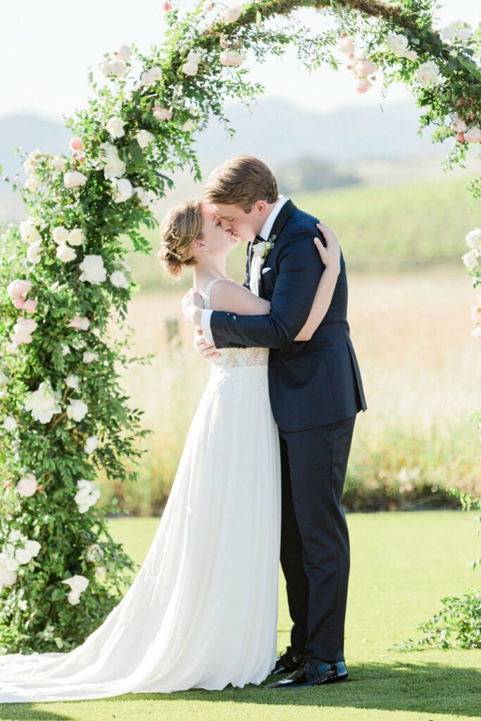 A bride in a white dress and a groom in a dark suit share a kiss under a floral arch outdoors.