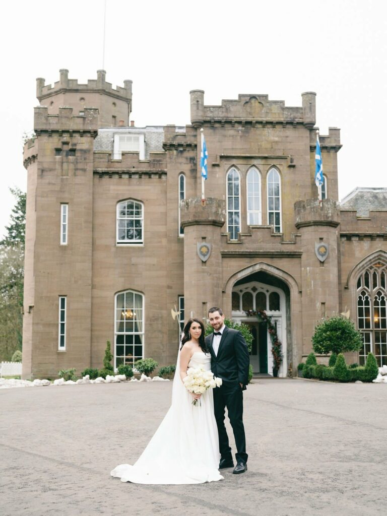 A bride and groom stand in front of a historic castle, with the bride holding a bouquet of flowers and the groom dressed in a black suit.