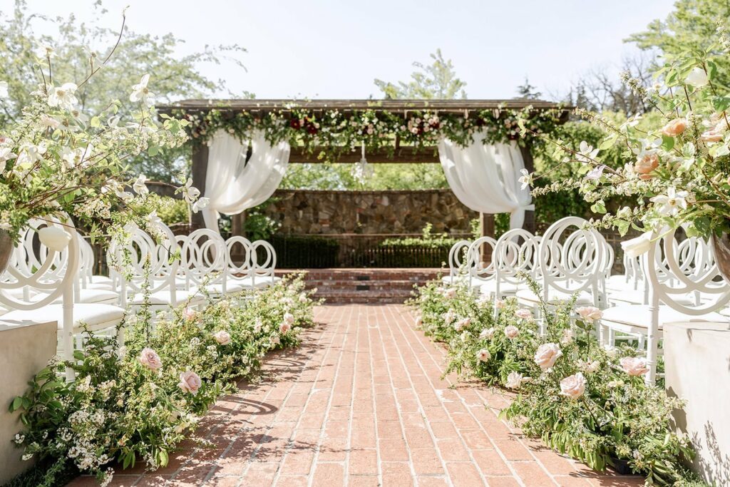 Outdoor wedding ceremony setup with white chairs on both sides of a brick aisle adorned with floral arrangements, leading to an altar covered with draped white fabric and greenery.