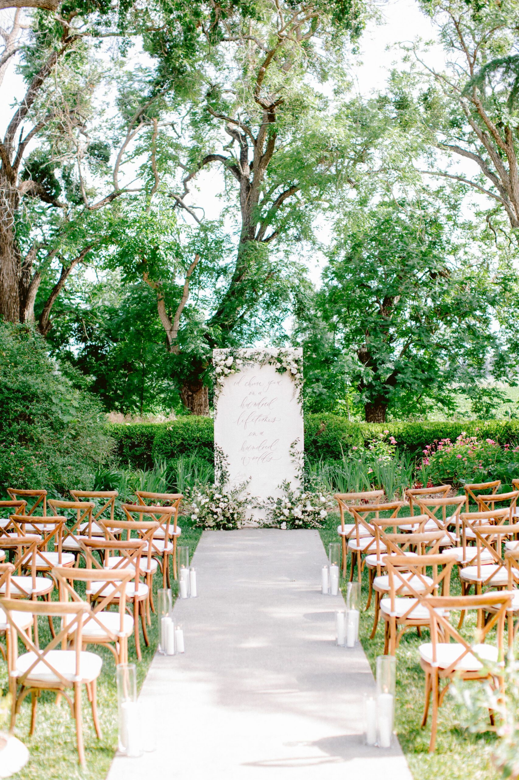 An outdoor wedding ceremony setup with rows of wooden chairs facing a floral-decorated arch and sign, bordered by trees and greenery. White candles line the aisle.