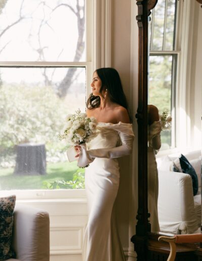 A woman in an off-the-shoulder white gown holds a bouquet while standing by a large window. She is looking outside and is reflected in a mirror beside her.