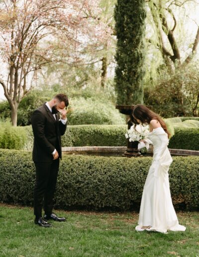 A bride in a white dress holding a bouquet and a groom in a black suit stand outside near a garden fountain. The groom covers his face with his hand while the bride looks down.