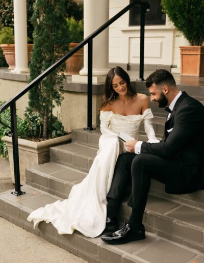 A bride in a white dress and a groom in a black suit sit on outdoor steps, reading together.