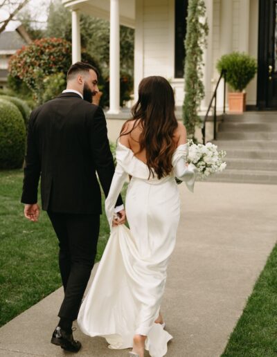 A bride and groom walk hand in hand toward a house. The bride is wearing a white off-shoulder gown and holding a bouquet of white flowers. The groom is in a black suit.