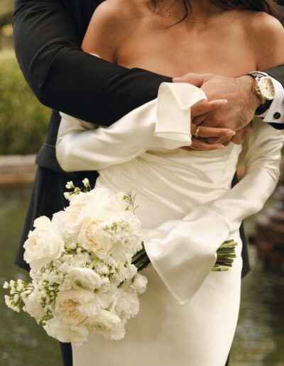 A person in a white wedding dress holding a bouquet of white flowers is embraced by another person in a dark suit with a gold watch.