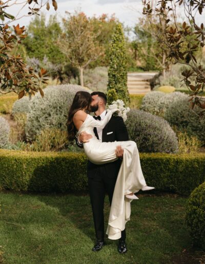 A groom in a black suit lifts and kisses a bride in a white gown in a manicured garden with tall hedges and greenery in the background.