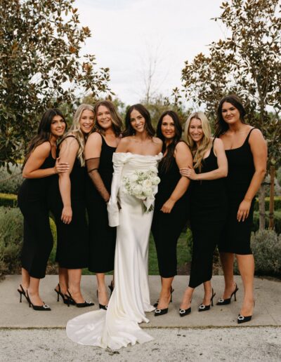 Bride in a white gown stands with six bridesmaids wearing black dresses and heels in an outdoor setting, flanked by large trees.