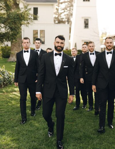 A group of men in black tuxedos with bow ties walk on a grass lawn with trees and white buildings in the background. The man in the center has a beard and is smiling slightly.