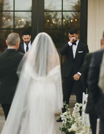 Bride walking down the aisle towards two men in suits, one of whom appears emotional and is wiping his eyes. The scene is outdoors in front of glass doors.