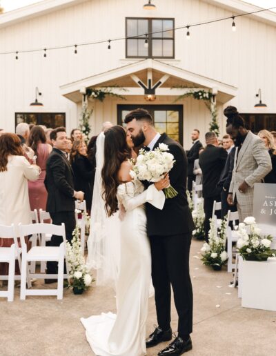 Bride and groom kiss in front of guests at an outdoor wedding ceremony near a white building, surrounded by flowers and decorations.