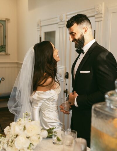 A bride and groom stand smiling at each other in a warmly lit room. The bride is in a white dress with a veil, and the groom is in a black tuxedo. A bouquet of white flowers is on the table beside them.