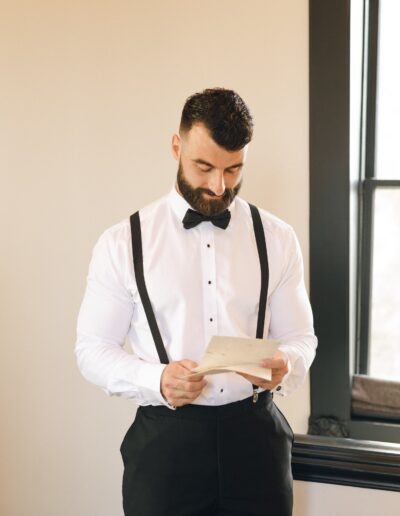 A bearded man dressed in a white shirt with black suspenders and a black bow tie stand indoors, looking down at a piece of paper in his hands.