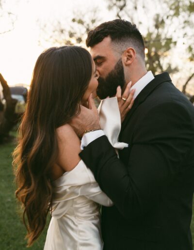 A bride in a white dress and a groom in a black suit share a kiss outdoors, with trees in the background.