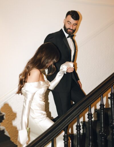 A couple dressed in formal attire descends a staircase; the woman is in an off-shoulder white dress, and the man in a black tuxedo.