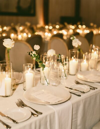 A long banquet table is set for an event with white plates, silverware, glassware, candles, and white floral centerpieces. The setting is softly lit, and rows of chairs line the table.