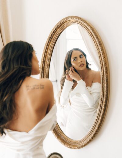 A woman in a white off-the-shoulder dress adjusts her earring while looking into an oval mirror. A tattoo is visible on her upper back.