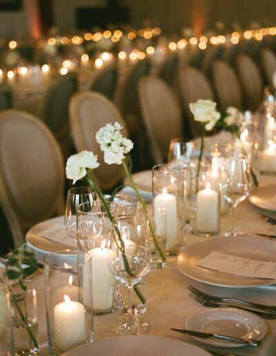 Elegant dining table set with white flowers, lit candles, wine glasses, plates, and menus, prepared for a formal event. Rows of chairs are visible in the background, with ambient lighting.