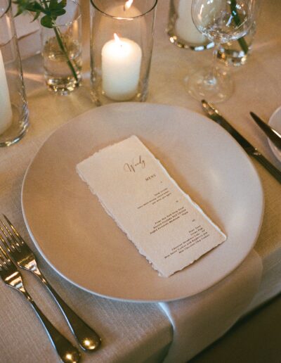 A white plate with a neatly placed menu on it, flanked by a fork and knife. Candles and wine glasses are arranged around the plate on a beige tablecloth.