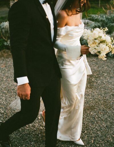 A bride in a white gown and groom in a black suit walk together on a rocky path; the bride holds a bouquet of white flowers.