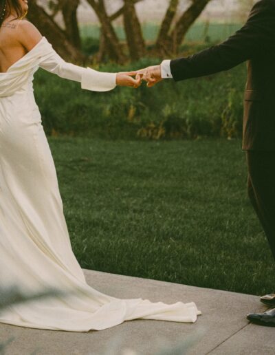 A bride in an off-the-shoulder white gown holds hands with a groom in a dark suit while walking outdoors on a grassy area.