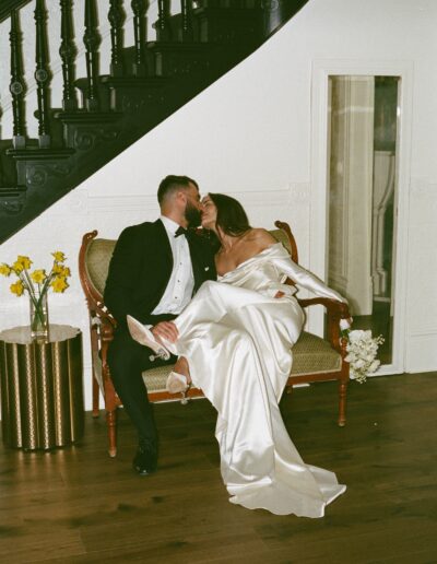 A couple in formal attire shares a kiss while seated on a vintage bench in front of a staircase. The bride holds a bouquet of flowers and is wearing a long, white satin dress.