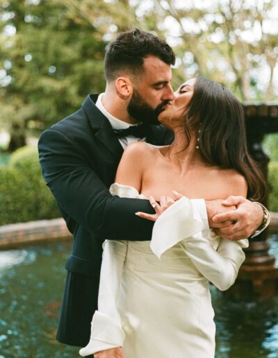 A couple in formal attire kisses in front of a fountain in a garden setting. The man wears a black suit and the woman wears a white off-shoulder dress.