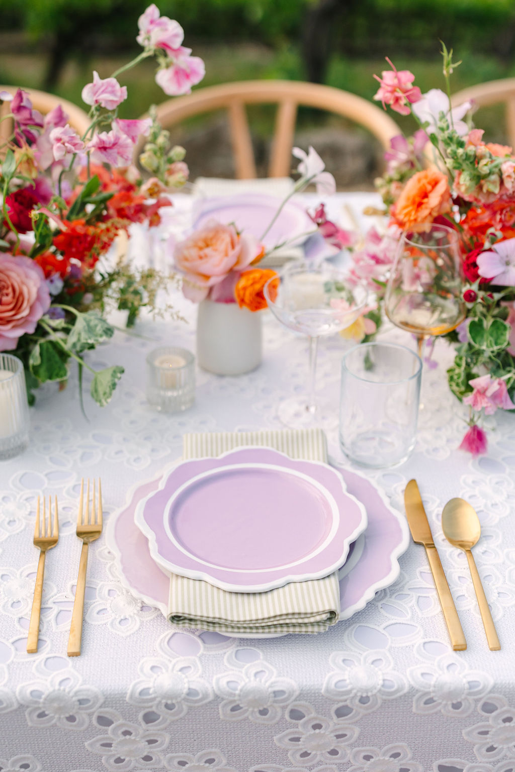 A table setting with purple plates, gold cutlery, and flower arrangements on a white lace tablecloth.