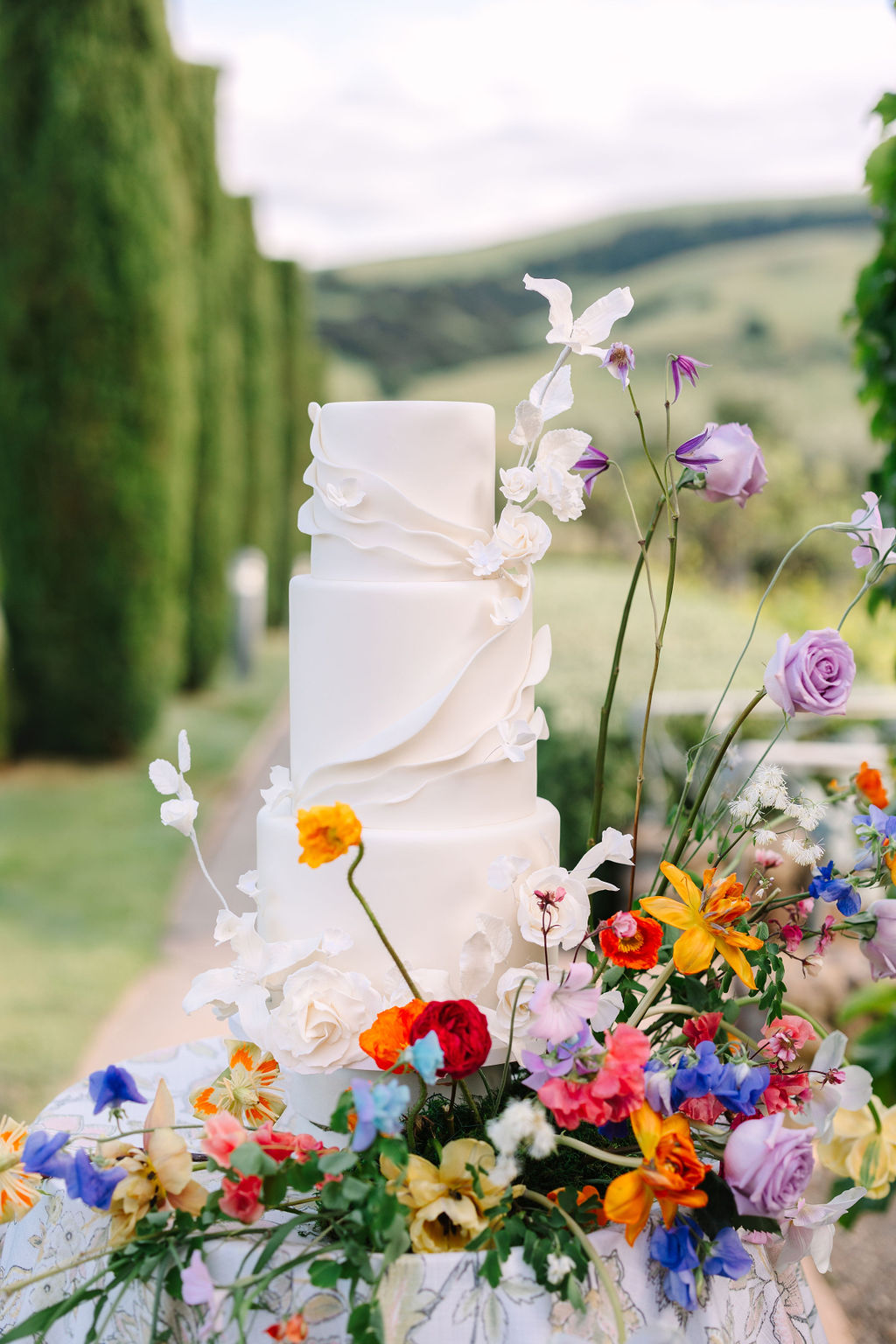 A four-tiered white wedding cake with delicate floral decorations is displayed outdoors, surrounded by colorful flowers. Lush green trees and a scenic backdrop are visible in the background.