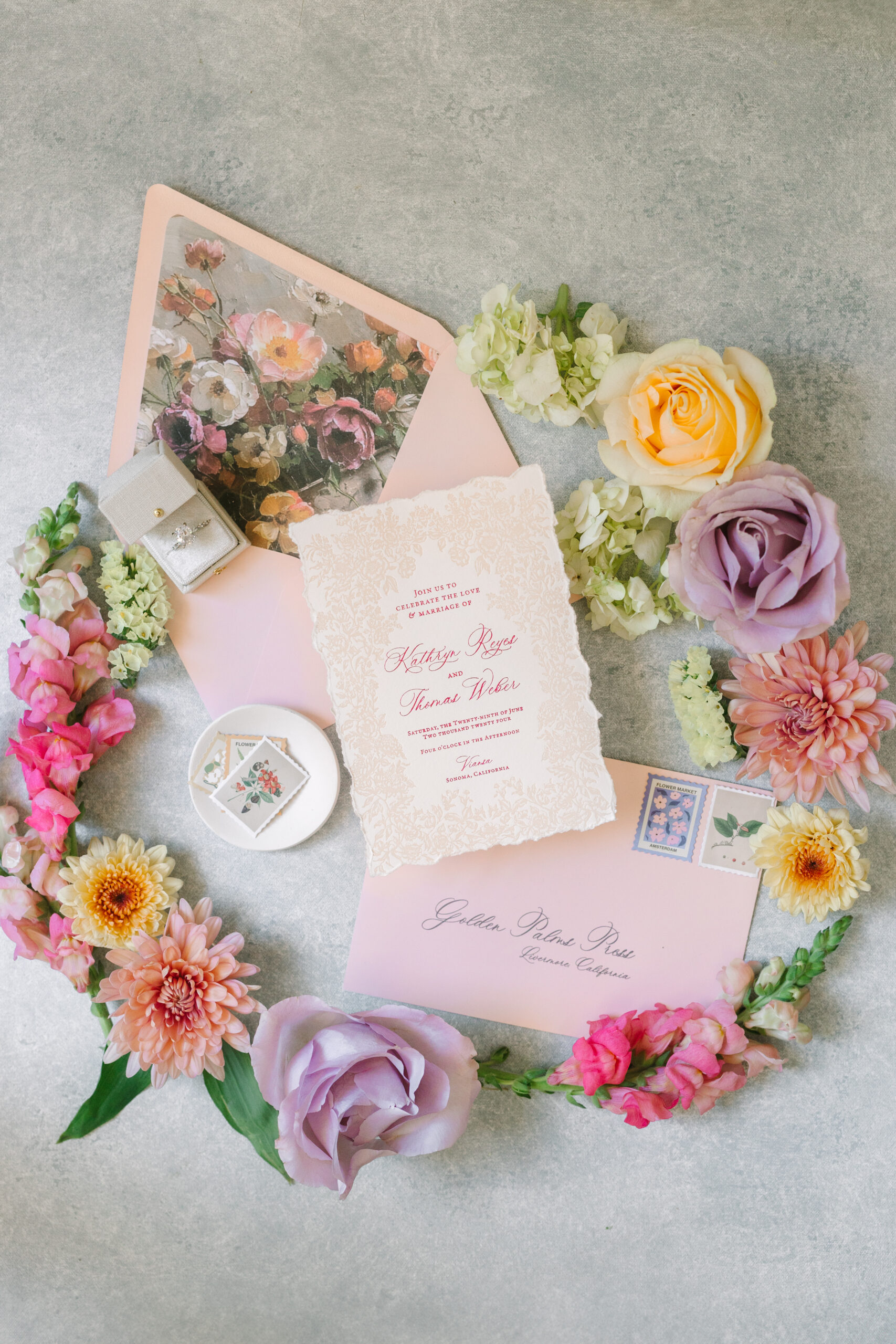 A wedding invitation set displayed with floral arrangements, stamps, a ring box, and envelopes on a light-colored surface.