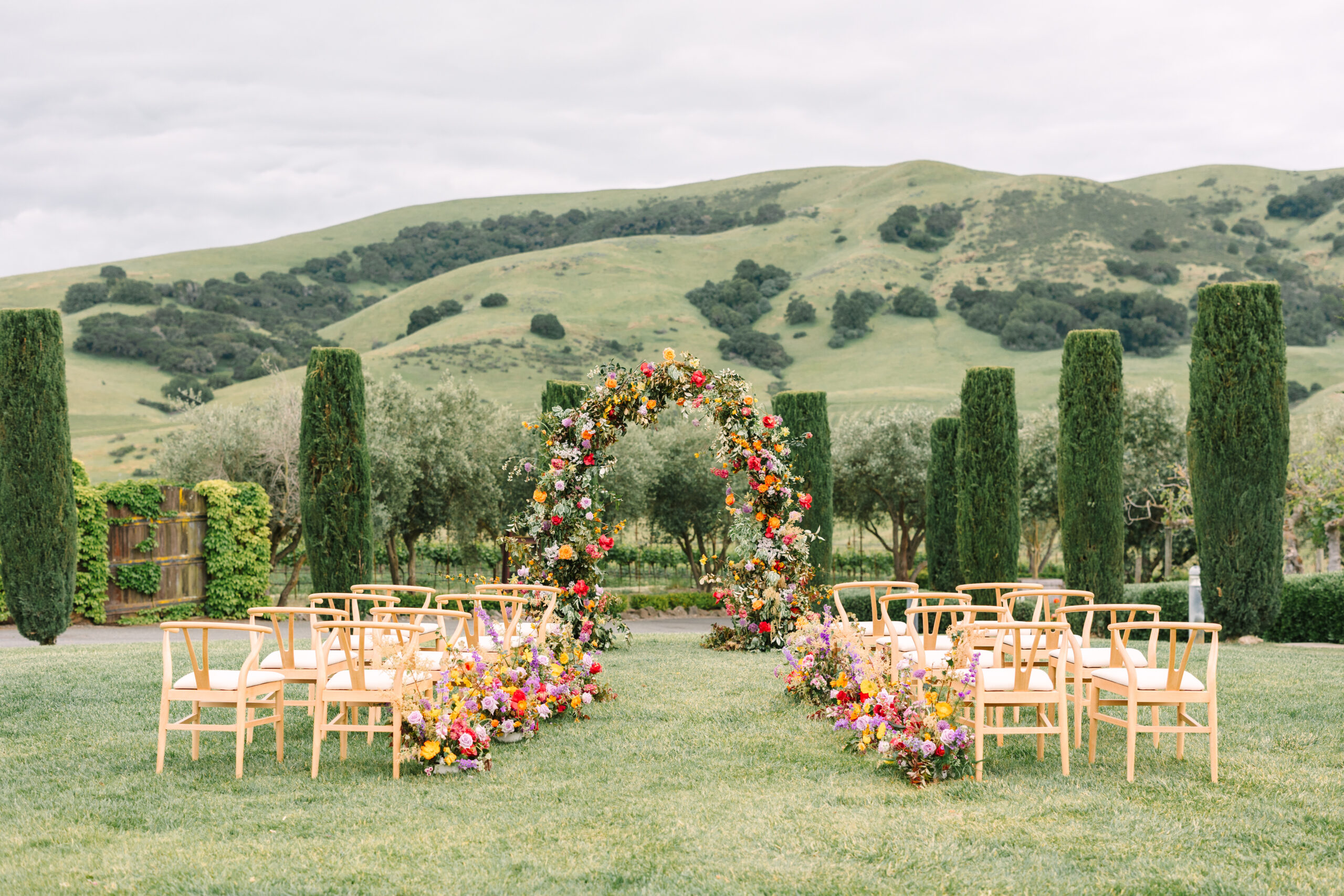 An outdoor wedding setup with wooden chairs arranged around a circular floral arch. The background features green hills and cypress trees.