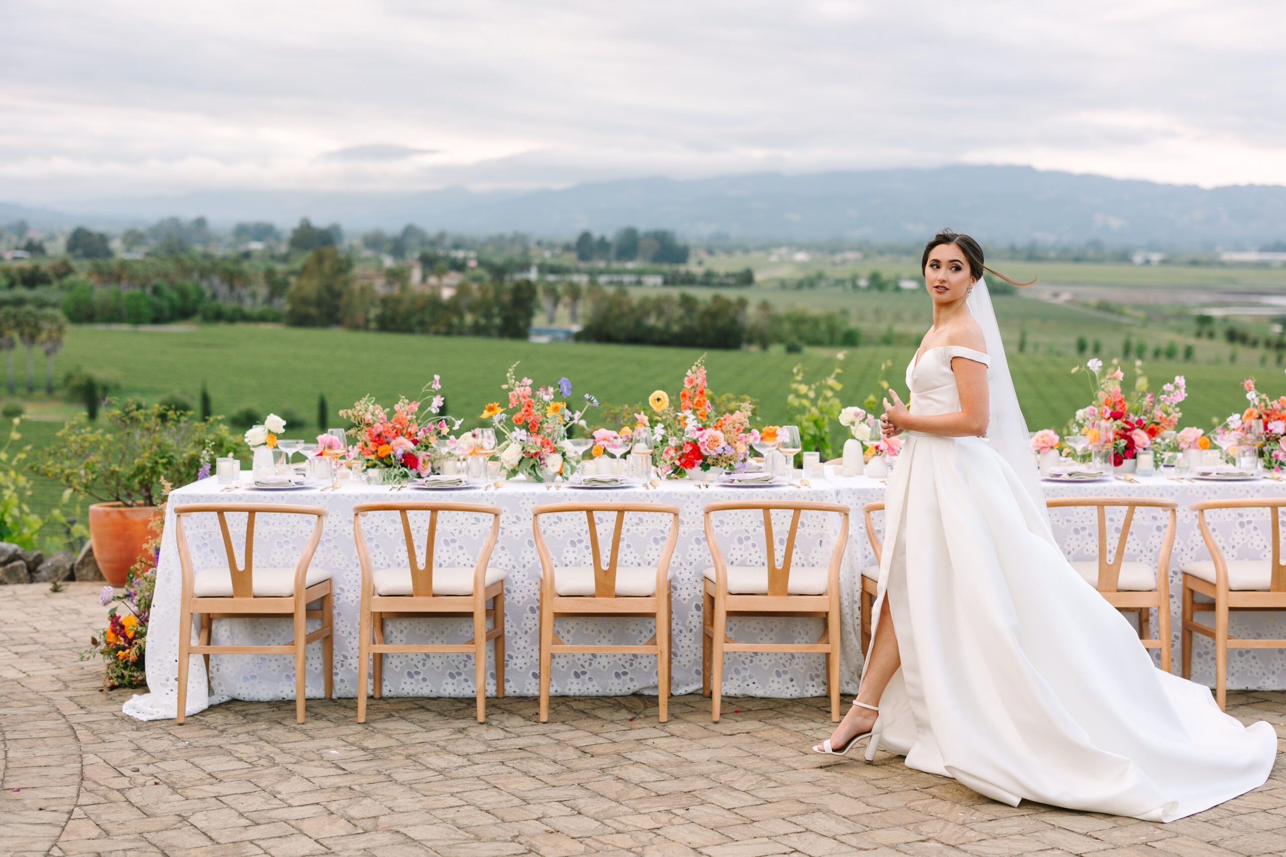 A bride in a white wedding dress stands by a long, decorated outdoor dining table, with a scenic view of lush fields and hills in the background.