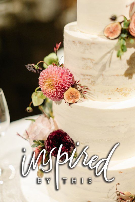 A three-tiered white wedding cake decorated with fresh pink and deep red flowers, slices of figs, and greenery. The words "inspired by this" are overlayed on the image.