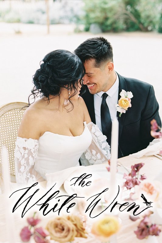 A bride and groom sit closely, smiling at each other. The bride wears an off-shoulder white dress, and the groom is in a black suit with a peach boutonniere. Text on the image reads "The White Wren.